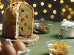 Un panettone già tagliato, senza una fetta, poggiato su un tagliere in legno e dietro di lui uno sfondo di una casa addobbata per il Natale