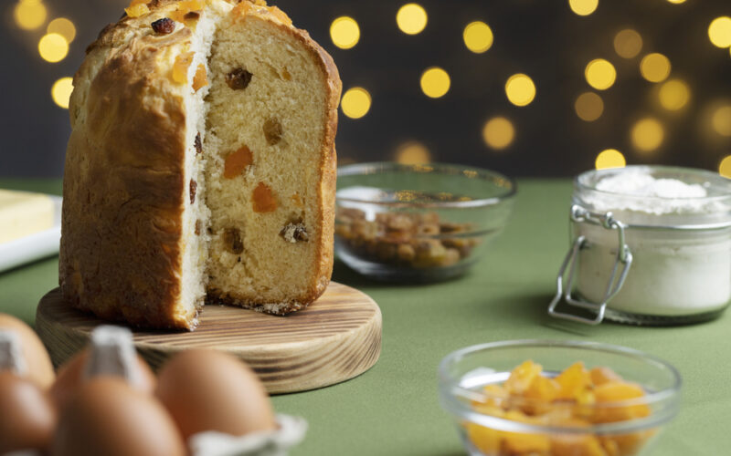 Un panettone già tagliato, senza una fetta, poggiato su un tagliere in legno e dietro di lui uno sfondo di una casa addobbata per il Natale