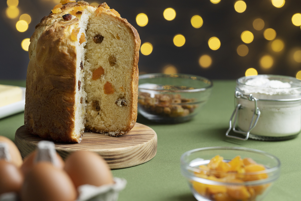 Un panettone già tagliato, senza una fetta, poggiato su un tagliere in legno e dietro di lui uno sfondo di una casa addobbata per il Natale