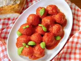 Un piatto di polpette al sugo decorate con delle foglie di basilico. Il piatto è messo su una tavola coperta da una tovaglia a quadretti bianchi e rossi.