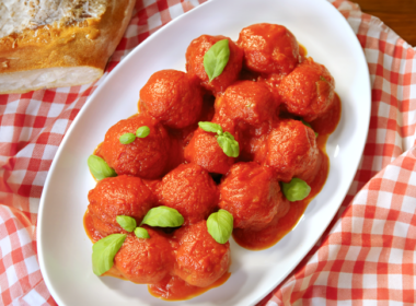 Un piatto di polpette al sugo decorate con delle foglie di basilico. Il piatto è messo su una tavola coperta da una tovaglia a quadretti bianchi e rossi.