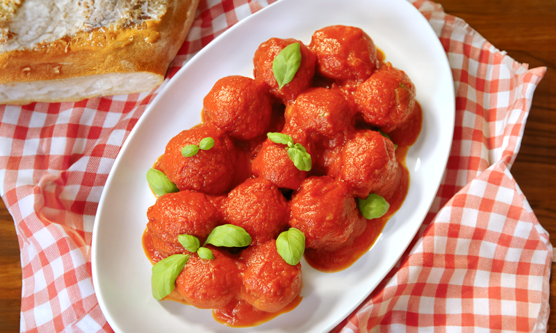 Un piatto di polpette al sugo decorate con delle foglie di basilico. Il piatto è messo su una tavola coperta da una tovaglia a quadretti bianchi e rossi.