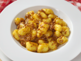 Un piatto fondo bianco pieno di gnocchi, poggiato su un altro piatto piano. Entrambi i piatti sono su un tavolo coperto da una tovaglia a quadretti bianchi e rosa