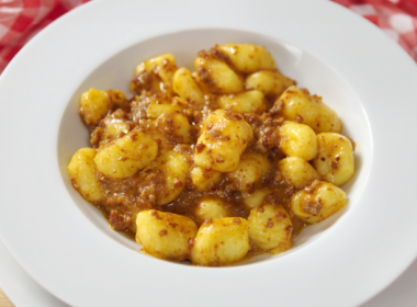 Un piatto fondo bianco pieno di gnocchi, poggiato su un altro piatto piano. Entrambi i piatti sono su un tavolo coperto da una tovaglia a quadretti bianchi e rosa