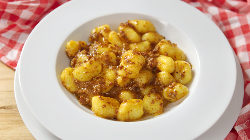 Un piatto fondo bianco pieno di gnocchi, poggiato su un altro piatto piano. Entrambi i piatti sono su un tavolo coperto da una tovaglia a quadretti bianchi e rosa