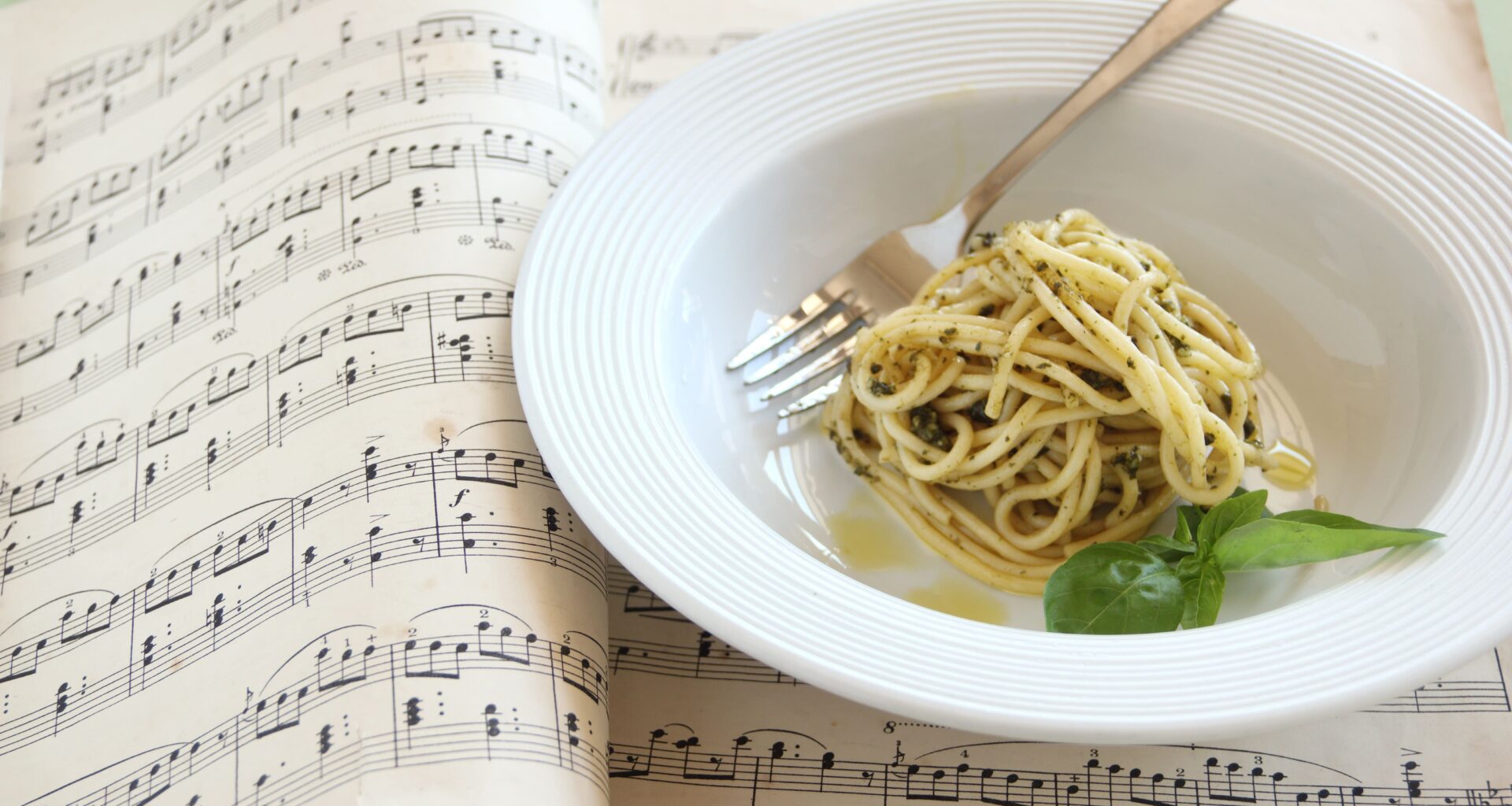 un piatto di spaghetti al pesto è poggiato su un quaderno di musica, in particolare su due pagine che contengono degli spartiti musicali. è una metafora del cibo nella musica