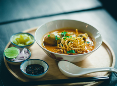 un vassoio in legno contiene i piatti che compongono una colazione salata. C'è una ciotola piena di una zuppa di noodles e altri condimenti accanto.