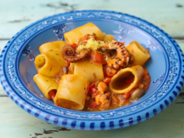 un piatto blu che contiene dei paccheri conditi con un sugo a base di pomodoro e polpo