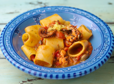 un piatto blu che contiene dei paccheri conditi con un sugo a base di pomodoro e polpo