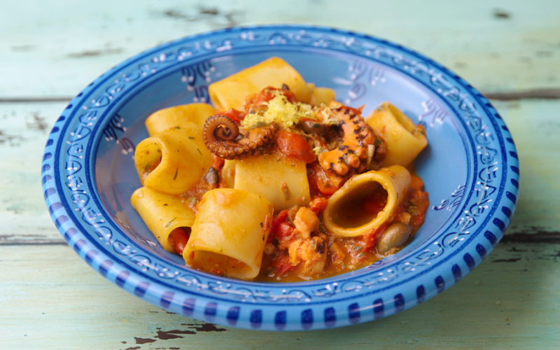 un piatto blu che contiene dei paccheri conditi con un sugo a base di pomodoro e polpo