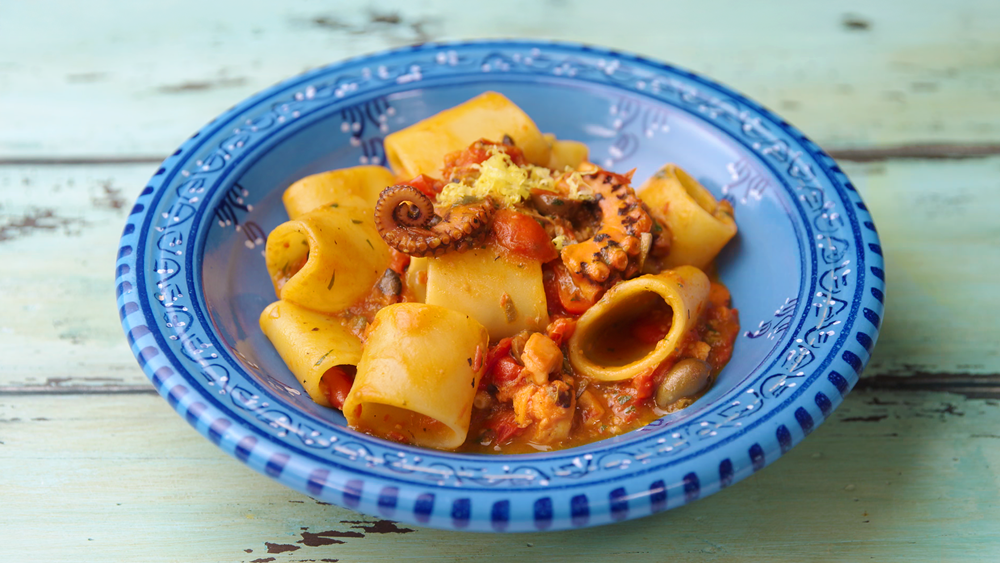 un piatto blu che contiene dei paccheri conditi con un sugo a base di pomodoro e polpo