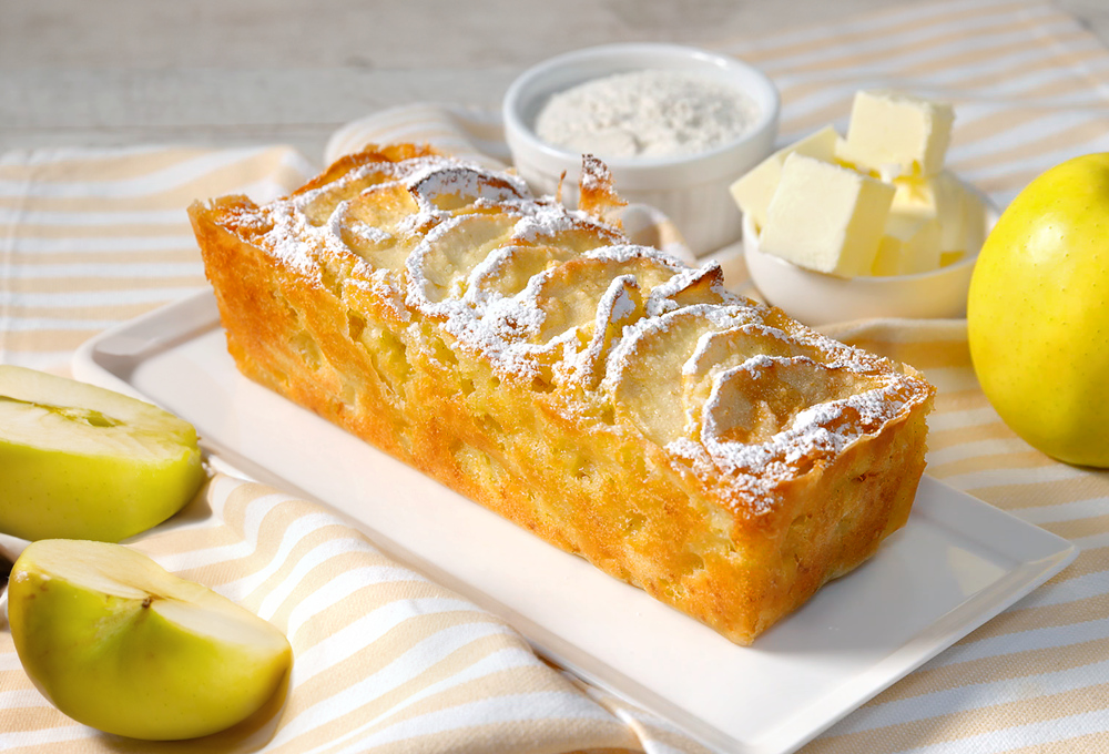 un plumcake alle mele è poggiato su piatto da portata. a destra c'è una piccola ciotola piena di burro a tocchetti e un'altra con della farina. A sinistra del kiwi a fette.