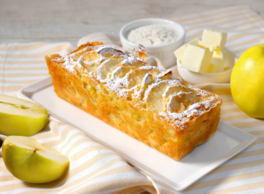 un plumcake alle mele è poggiato su piatto da portata. a destra c'è una piccola ciotola piena di burro a tocchetti e un'altra con della farina. A sinistra del kiwi a fette.