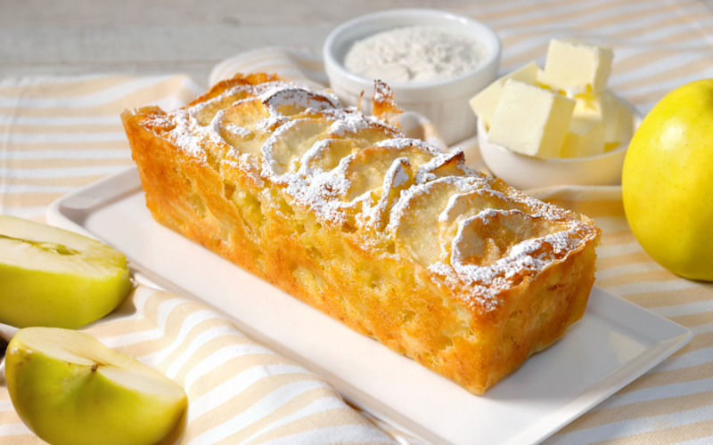 un plumcake alle mele è poggiato su piatto da portata. a destra c'è una piccola ciotola piena di burro a tocchetti e un'altra con della farina. A sinistra del kiwi a fette.