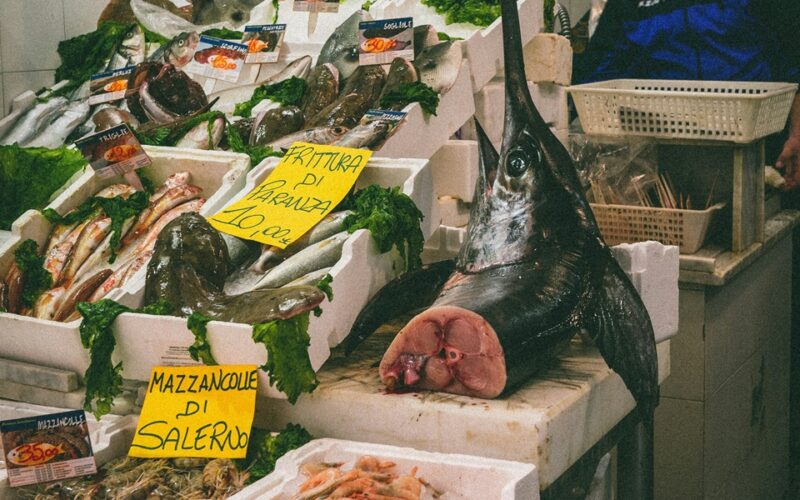 un frame di un mercato del pesce con in vendita un pezzo di pesce spada di cui si intravede l'interno e il prezzo