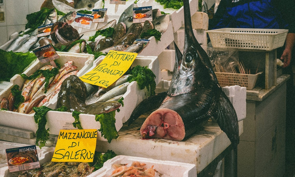 un frame di un mercato del pesce con in vendita un pezzo di pesce spada di cui si intravede l'interno e il prezzo