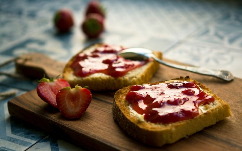 Un tagliere in legno su cui sono poggiate due fette di pane tostato su cui è stata spalmata della marmellata di fragole