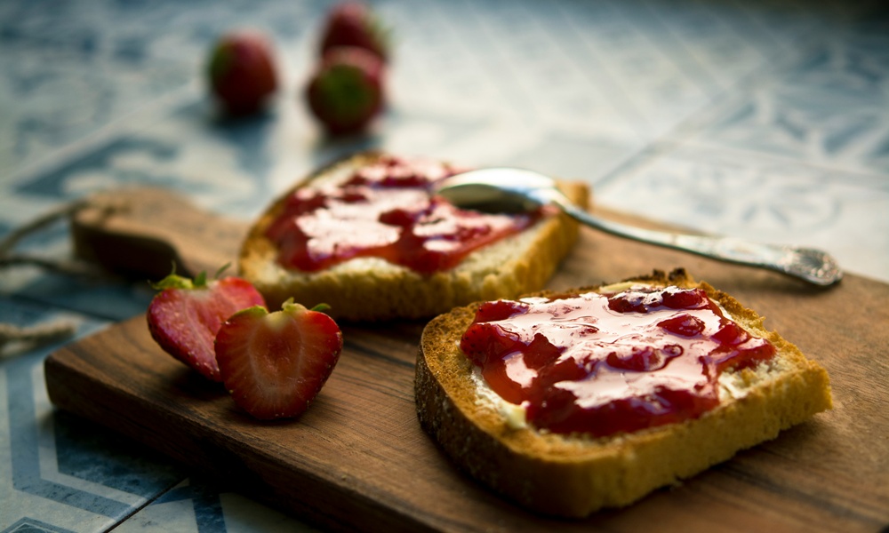 Un tagliere in legno su cui sono poggiate due fette di pane tostato su cui è stata spalmata della marmellata di fragole