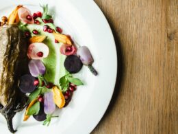 un piatto bianco che contiene un piatto a base di verdure,nello specifico una melanzana guarnita con fiori edibili, melograno e foglie di insalata