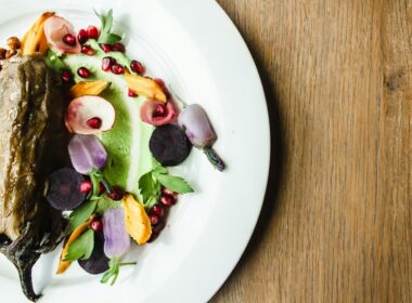 un piatto bianco che contiene un piatto a base di verdure,nello specifico una melanzana guarnita con fiori edibili, melograno e foglie di insalata