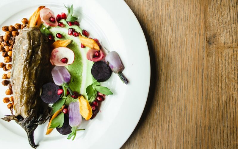 un piatto bianco che contiene un piatto a base di verdure,nello specifico una melanzana guarnita con fiori edibili, melograno e foglie di insalata