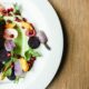un piatto bianco che contiene un piatto a base di verdure,nello specifico una melanzana guarnita con fiori edibili, melograno e foglie di insalata