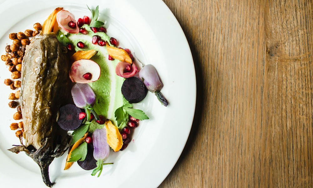 un piatto bianco che contiene un piatto a base di verdure,nello specifico una melanzana guarnita con fiori edibili, melograno e foglie di insalata