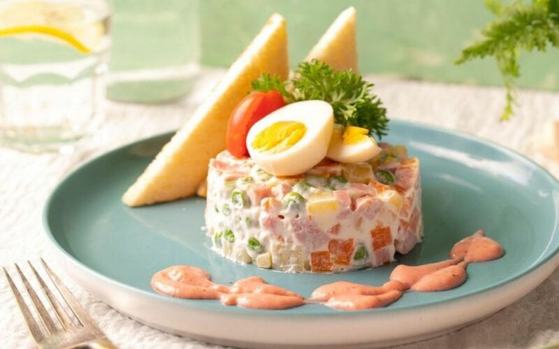 un piatto con una porzione di insalata russa guarnita con una salsa di accompagnamento e delle uova sode sopra